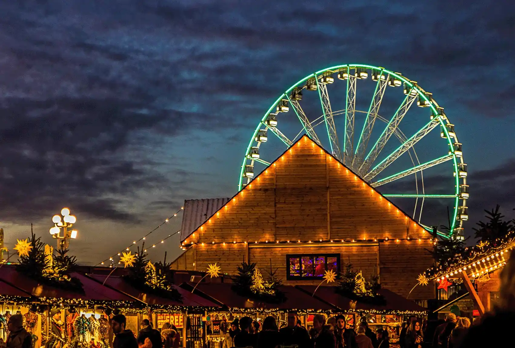 Winter Wonderland Hyde Park in London boasts an illuminated Ferris wheel, wooden stalls and twinkling Christmas decorations. Christmas market stalls under the glowing Ferris wheel.