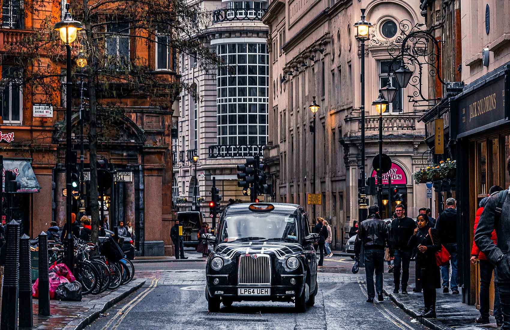 A distinctive scene of London: a black taxi driving through the city’s historic streets. Experience the charm of London’s old architecture Classic black London taxi driving through a historic street in the city.